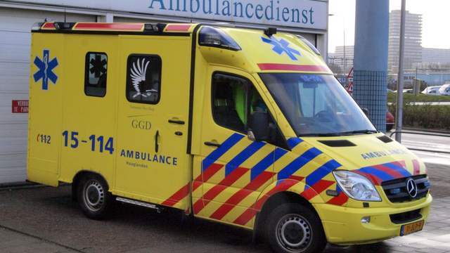 A yellow Dutch ambulance with blue and red stripes and the Dutch emergency number on its side, parked outside an ambulance station.