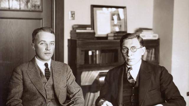 Two young men in suits sit side by side in an office, in front of a bookcase. A black-and-white photo.