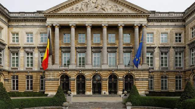 A pale stone building with tall columns, a carved front and two flags, behind a neat garden.