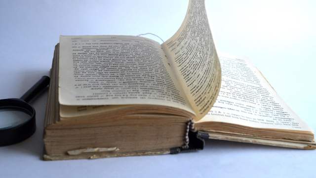 An old open book with one page lifting as it turns, and a magnifying glass lying beside it.