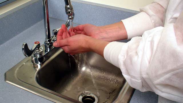 Two hands held under a running tap over a steel sink. The person wears a white protective gown; no face is shown.