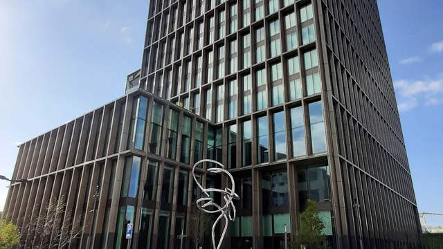 A tall office tower with a grid of dark windows under a blue sky, with a metal sculpture by the entrance.