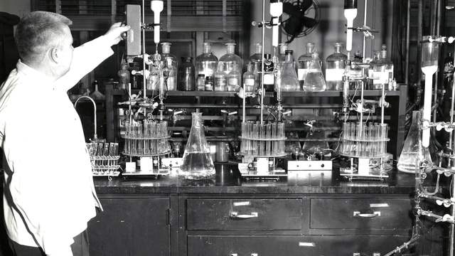 A man in a white shirt reaches up to a lamp in a laboratory full of glass flasks, bottles and racks of test tubes.