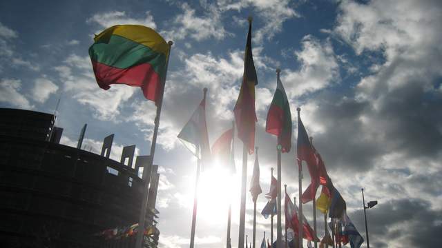 A row of national flags on tall poles in front of a round glass building, with the sun behind clouds.