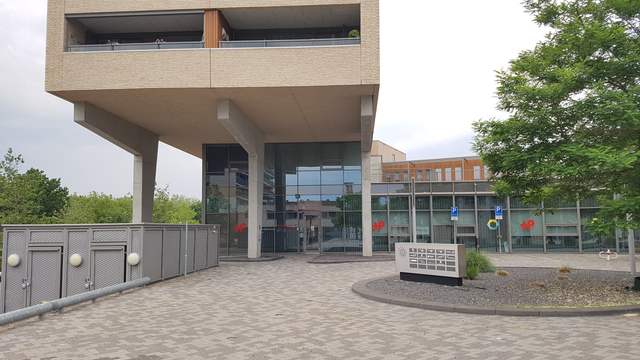The glass entrance of a modern Dutch health building under a large overhanging floor, with a tree and a paved square in front.
