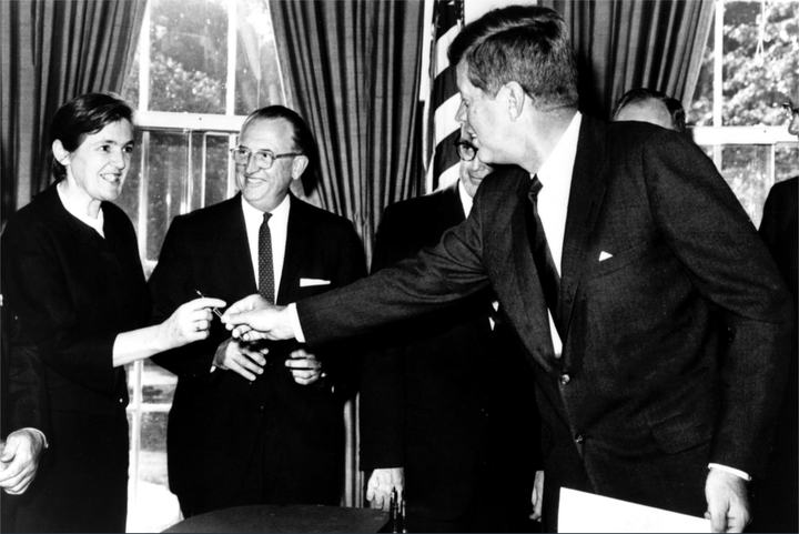 A black-and-white photo of President John F. Kennedy handing a pen to Frances Kelsey, with men in suits watching.