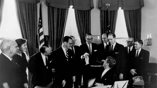 President Kennedy sits at his desk and hands a pen to a senator. Men in suits and one woman stand around him.