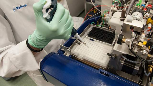 A gloved hand puts a small sample into a tray of wells on a large laboratory machine full of tubes and wires.