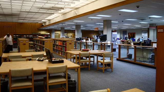 A large library reading room with wooden tables, chairs and low bookshelves, and computer desks further back.
