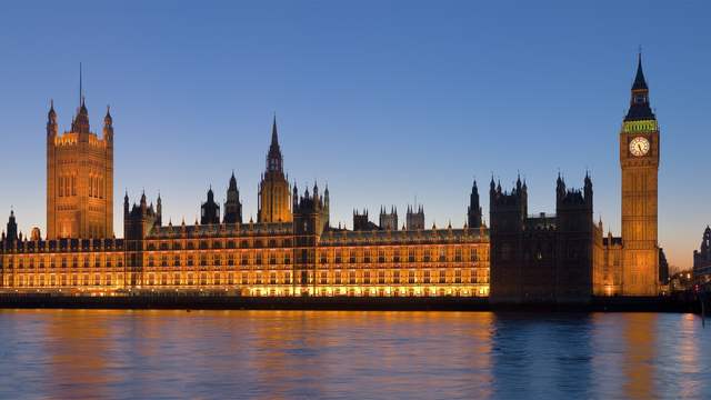 A long gothic palace with towers and a clock tower, lit up at dusk and reflected in a river.