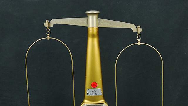 An old balance with two empty metal pans hanging from a gold-coloured stand on a wooden box with two drawers.