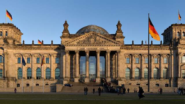 A large stone building with columns, four corner towers and a glass dome, with German flags, in evening light.