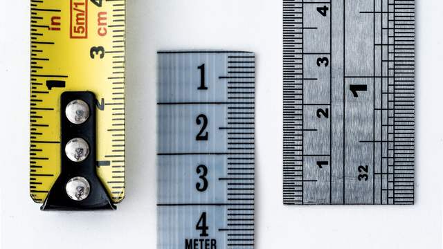 Three measuring scales side by side on a white surface: the end of a yellow tape measure, a pale blue tape marked in centimetres and a metal ruler, each marked differently.