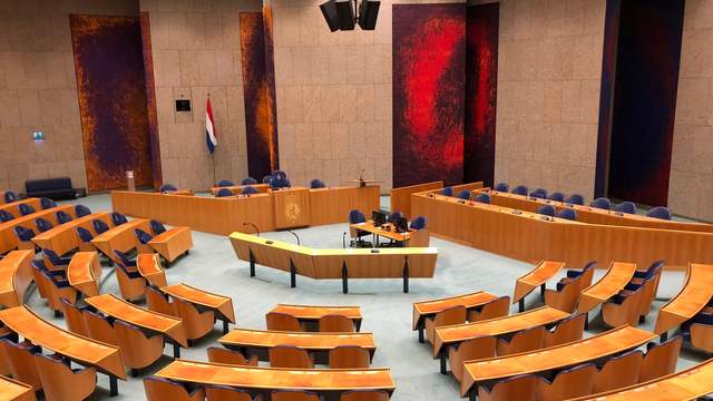 An empty debating chamber with rows of wooden desks and blue chairs in a half circle, a speaker's desk and a Dutch flag.