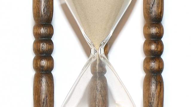 A wooden hourglass standing upright, with sand running from the top glass into the bottom glass.