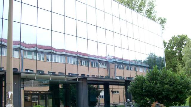 The main building of the School of Medicine in Zagreb: a glass front that reflects an older building, above a paved square with benches and a small statue.